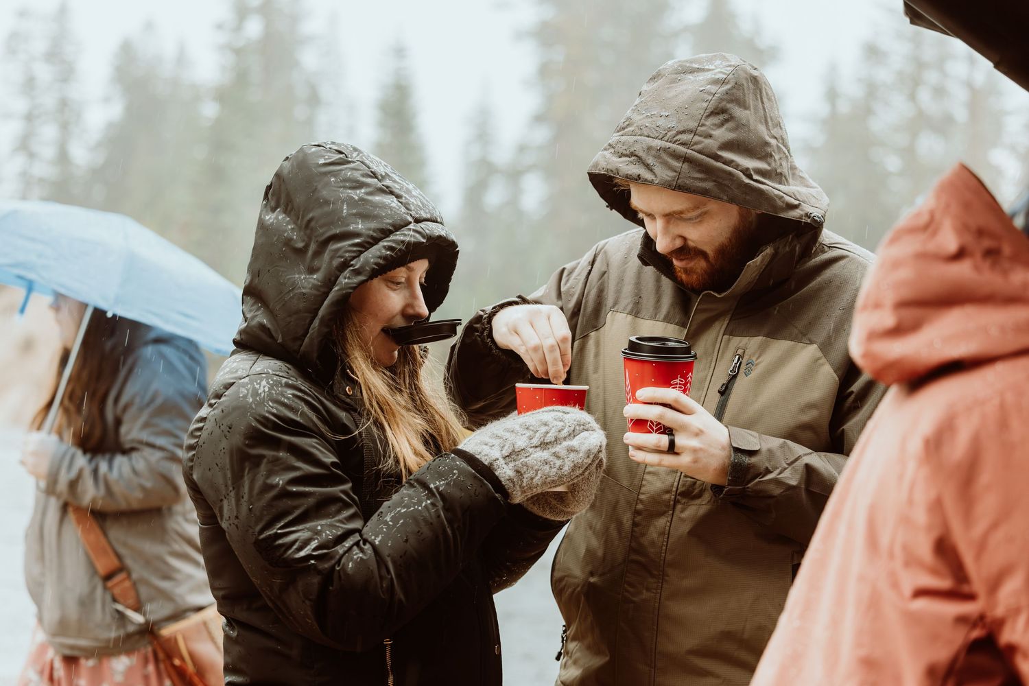Couple in rain jackets sharing hot coffee drinks outdoors in rainy forest weather.