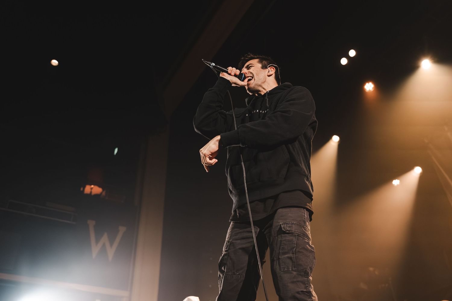 Male vocalist performing on stage holding microphone under golden stage lights at indoor music venue.