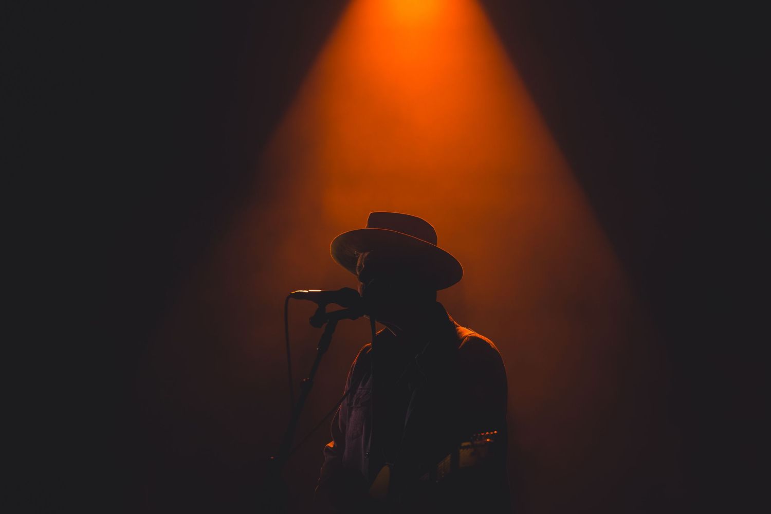 Singer in a wide-brim hat performing at a microphone under a warm orange stage spotlight.