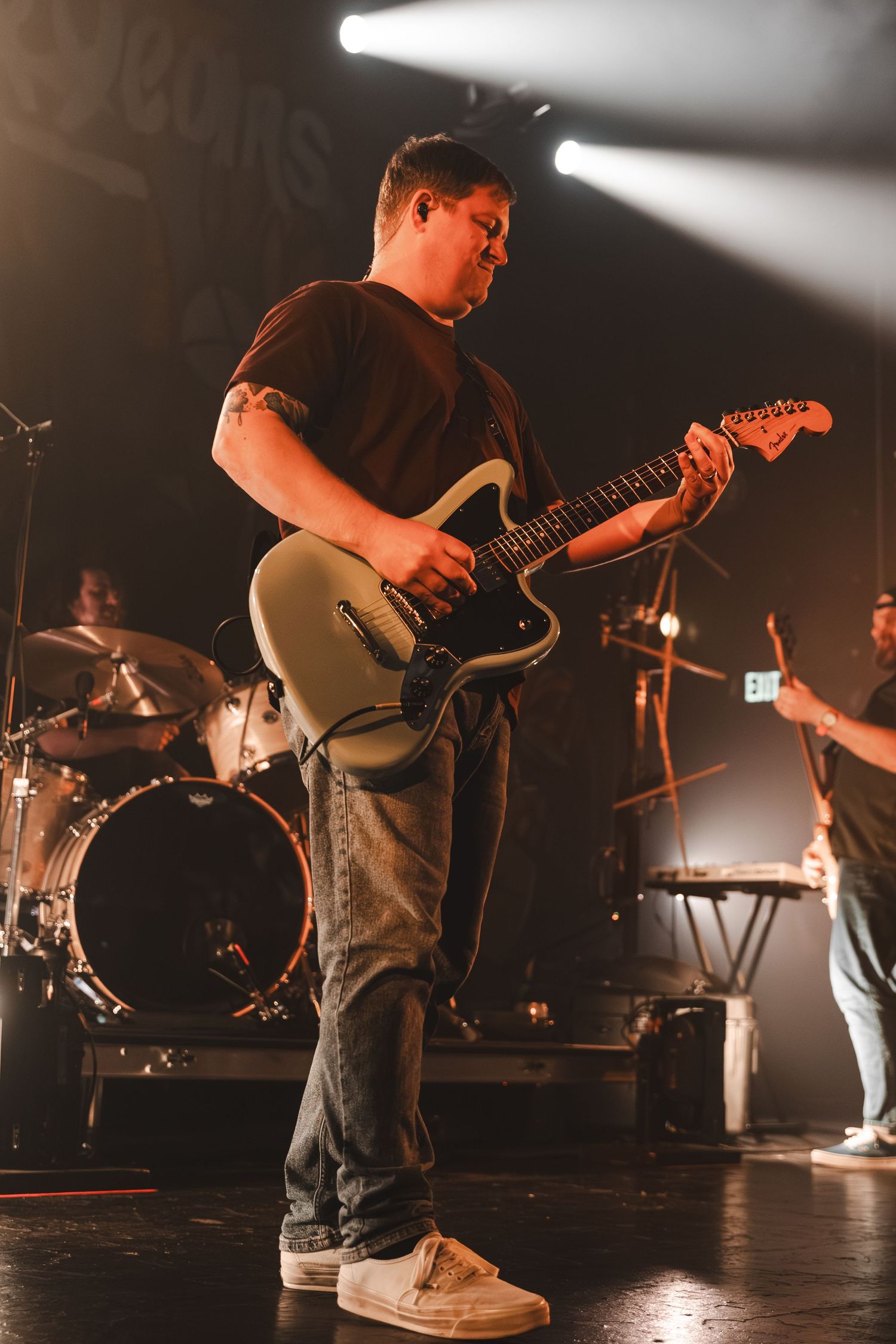 Guitarist plays silver Fender guitar on stage with drum kit visible in background at concert.