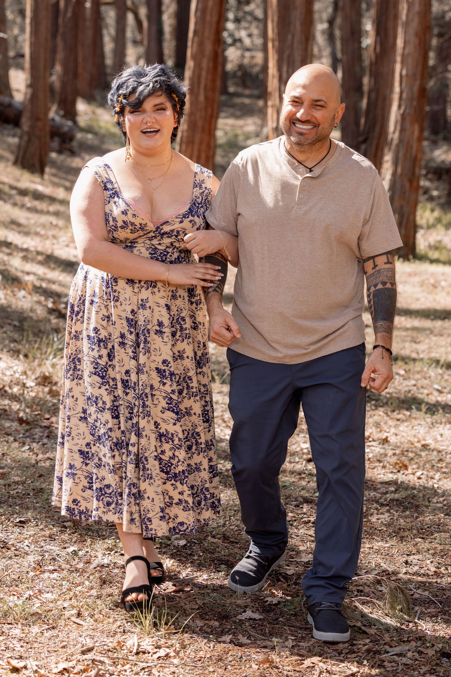 Happy couple walking hand-in-hand through sun-dappled pine forest, woman in floral midi dress.