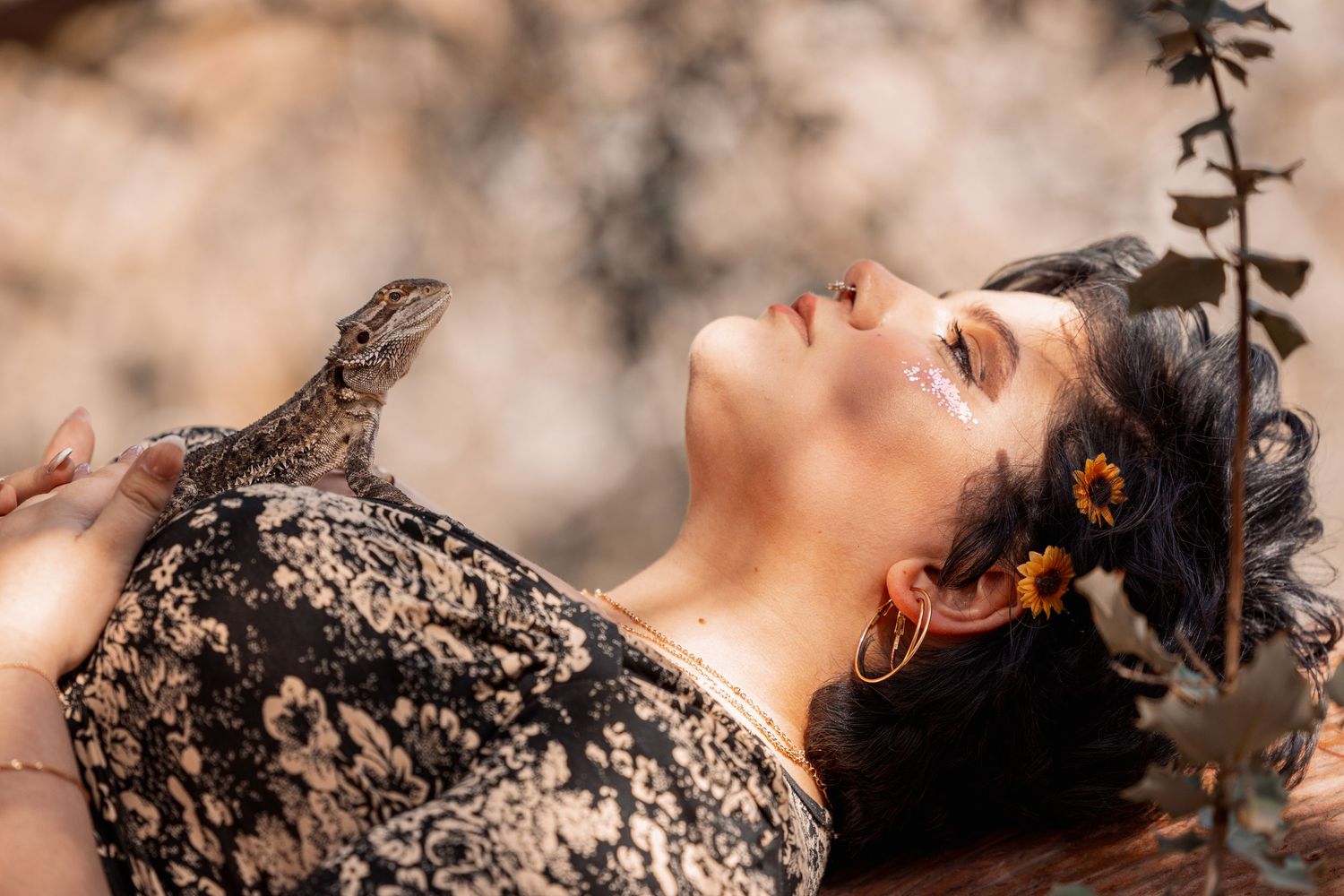 Woman with sunflower hair accessories reclines outdoors with a lizard resting on her chest.