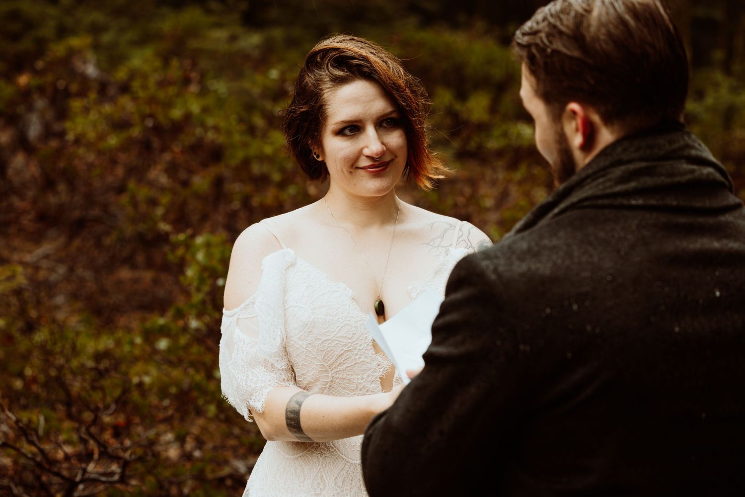 Bride smiles warmly at groom during outdoor forest wedding ceremony surrounded by lush greenery.