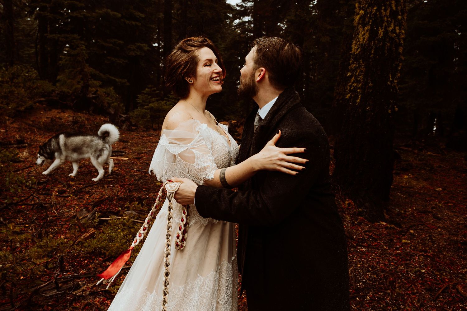 Couple embracing and laughing with their husky dog in the background during forest wedding.