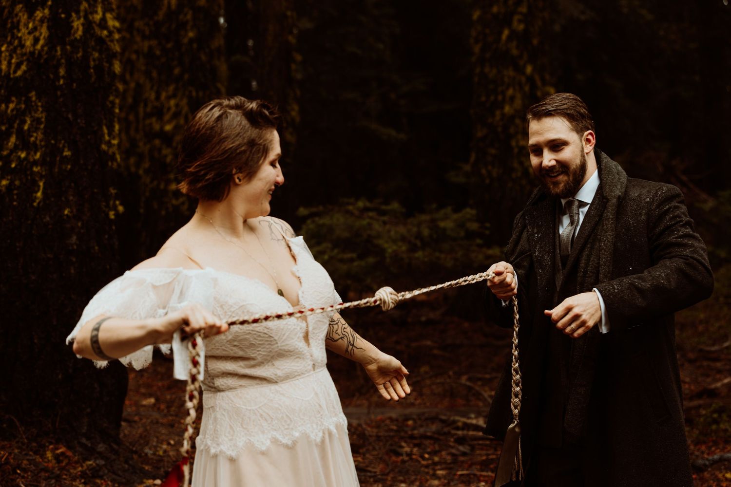 Bride and groom laughing while holding a handfasting cord during their forest elopement.