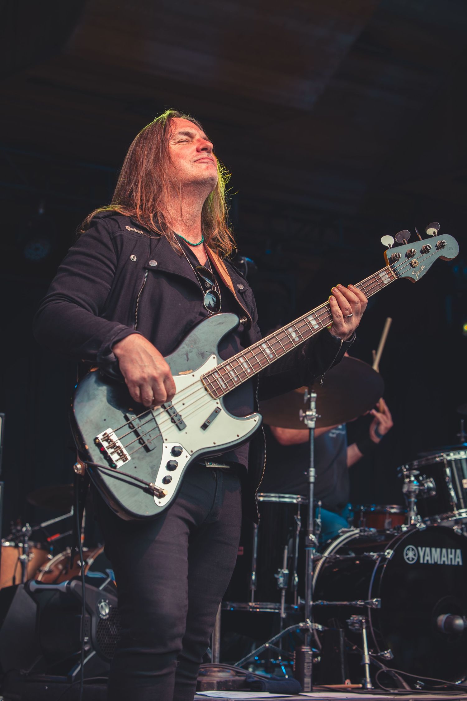 Long-haired bassist playing a blue electric guitar on a lit concert stage.