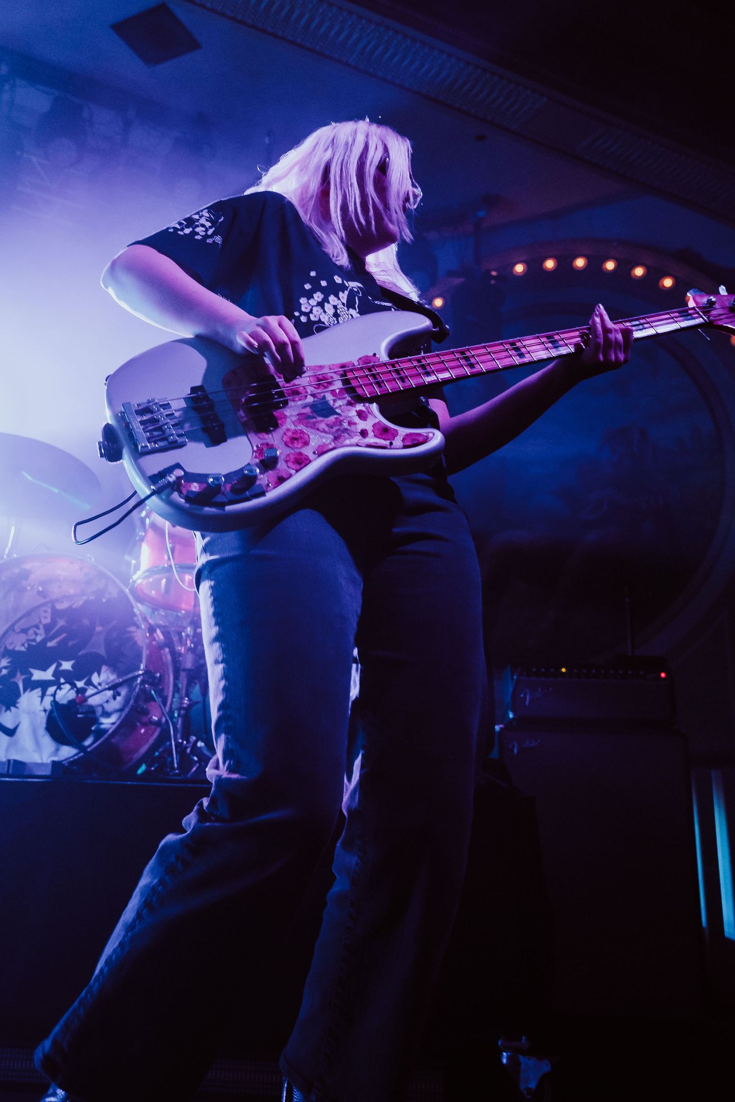 Female bassist with blonde hair playing a floral guitar under purple stage lights.