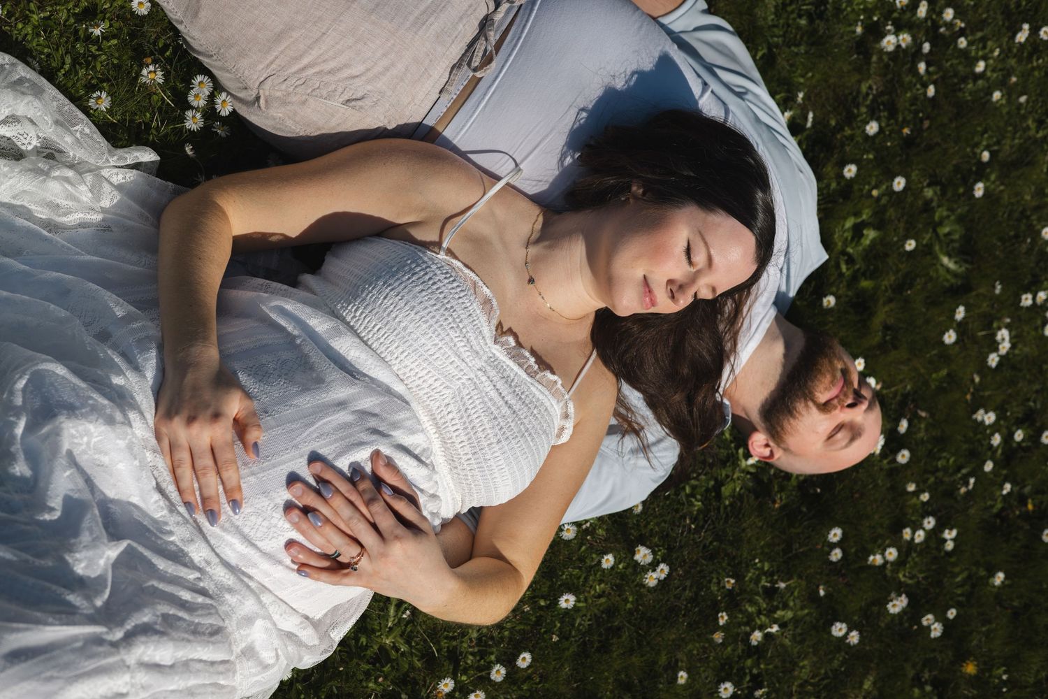 Couple lying together in a daisy field, woman in white dress resting peacefully on a sunny day.