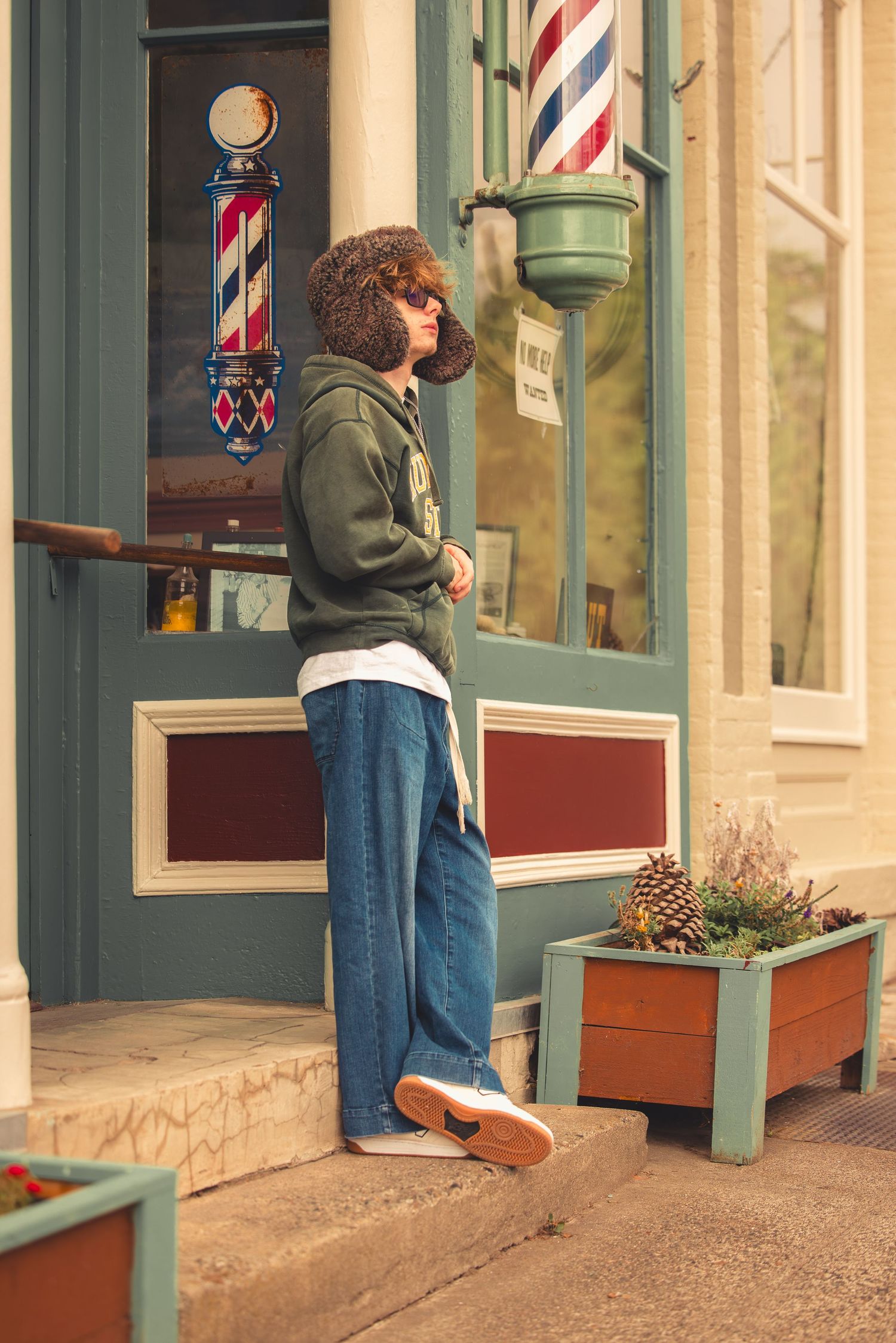 Curly-haired teen in green hoodie and wide-leg jeans stands outside vintage barbershop storefront.