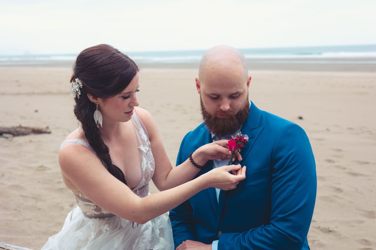 Bride carefully attaches floral boutonniere to groom's blue suit lapel on sandy beach.