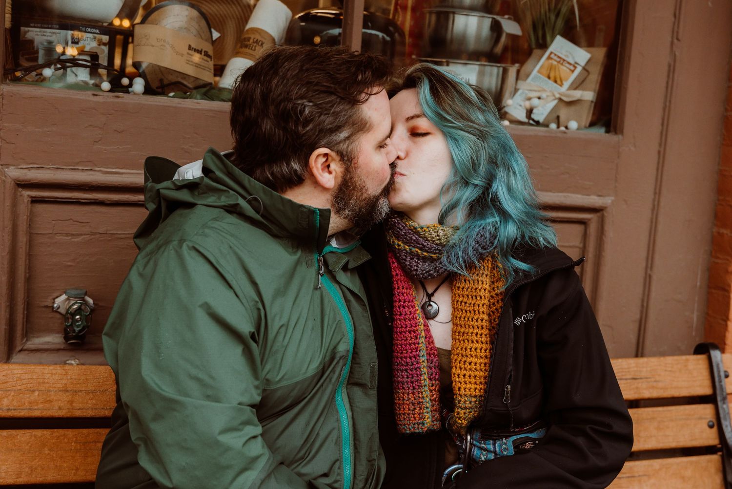 Couple kissing on a bench outside a rustic storefront, woman wearing a colorful knit scarf.
