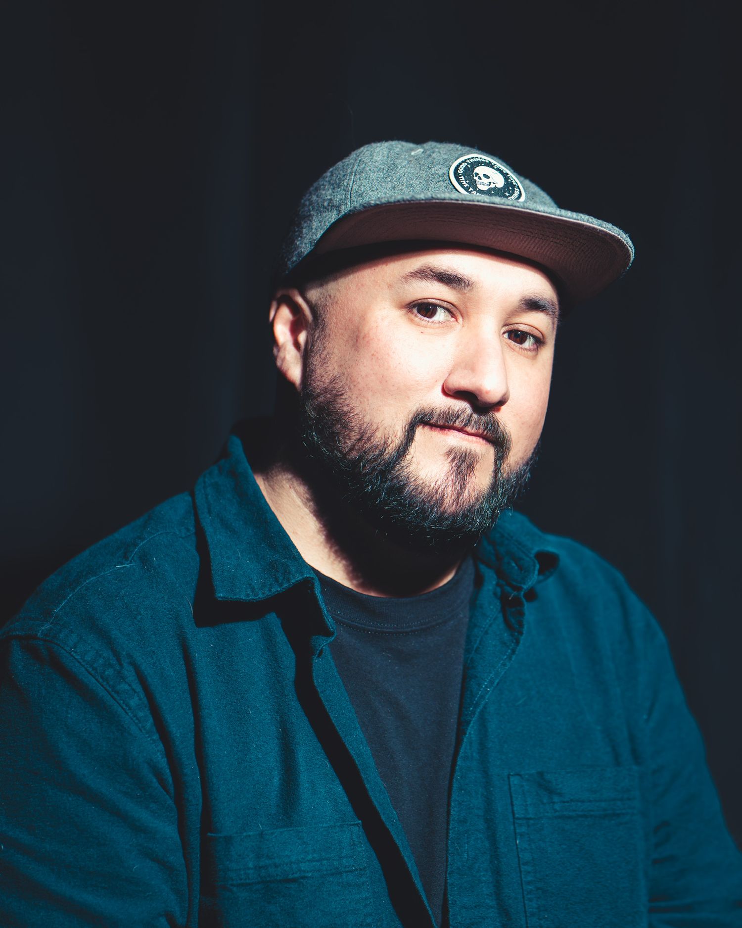 A bearded man wearing a gray snapback cap and blue denim shirt poses for a portrait against a dark background.