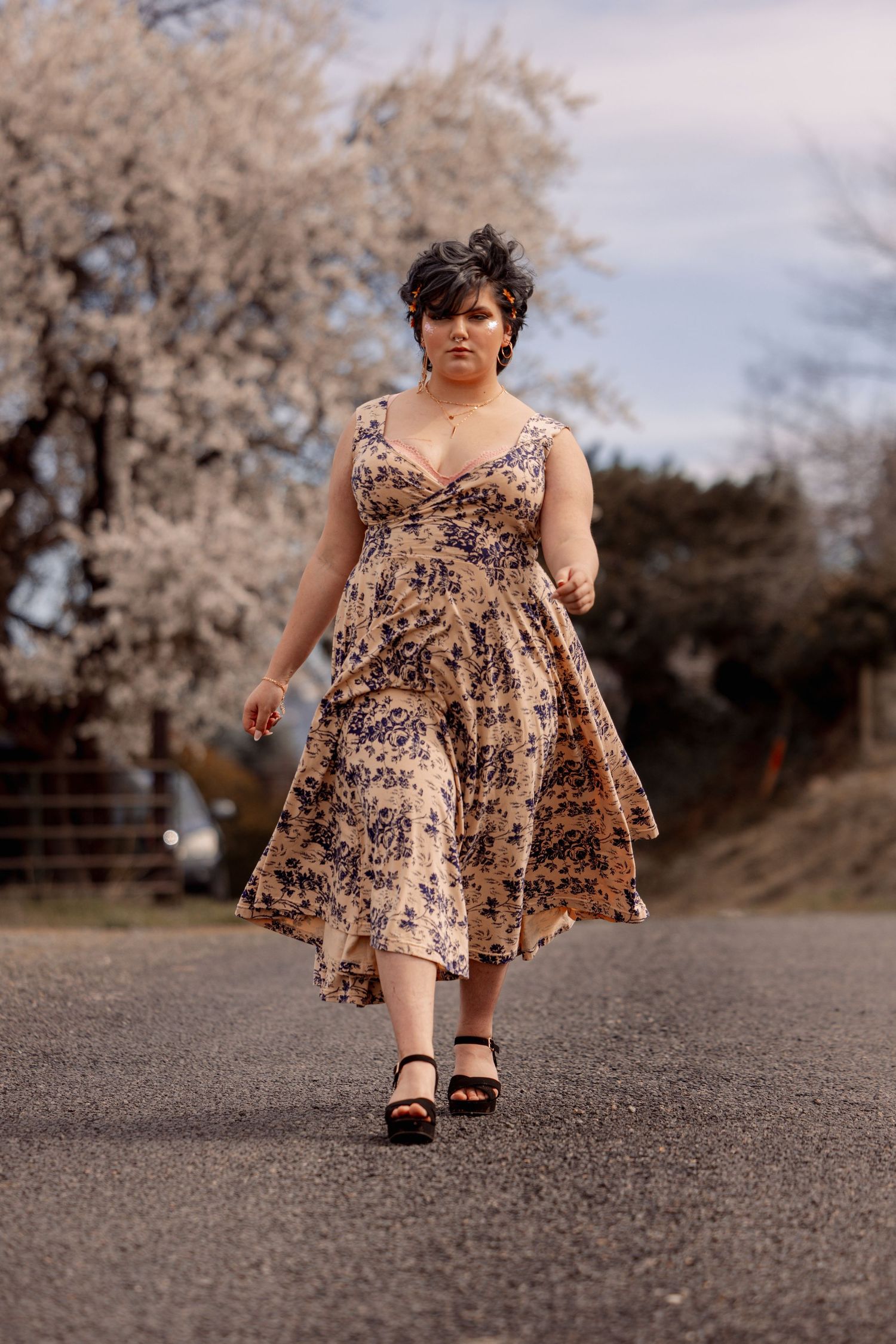 Woman walking confidently in floral midi dress on rural road with blooming trees behind her.