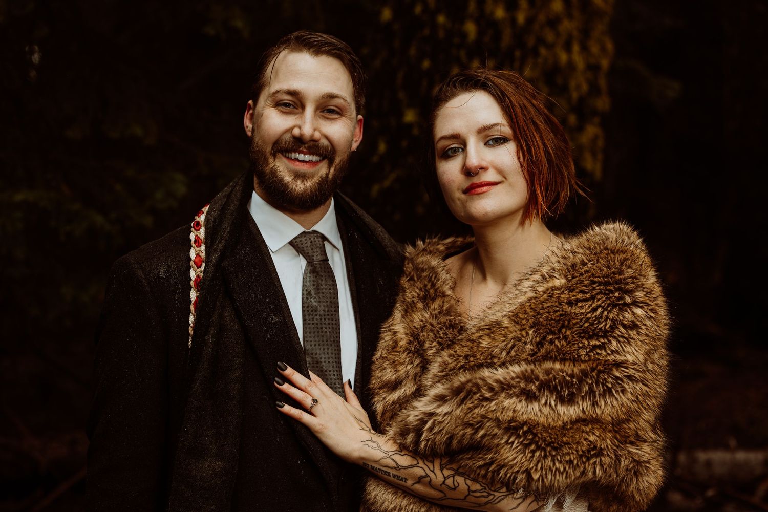 Smiling bride and groom posing in a dark forest, bride wearing a brown fur wrap wedding portrait.