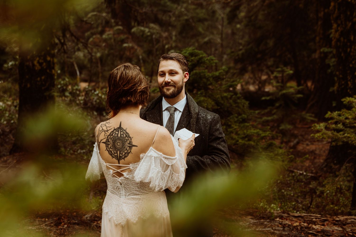 Smiling groom reads vows to bride with back tattoo during a dreamy forest wedding ceremony.