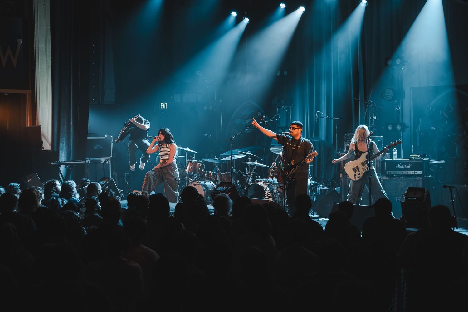 Full band performs on stage with dramatic blue and white spotlights illuminating silhouetted crowd below.