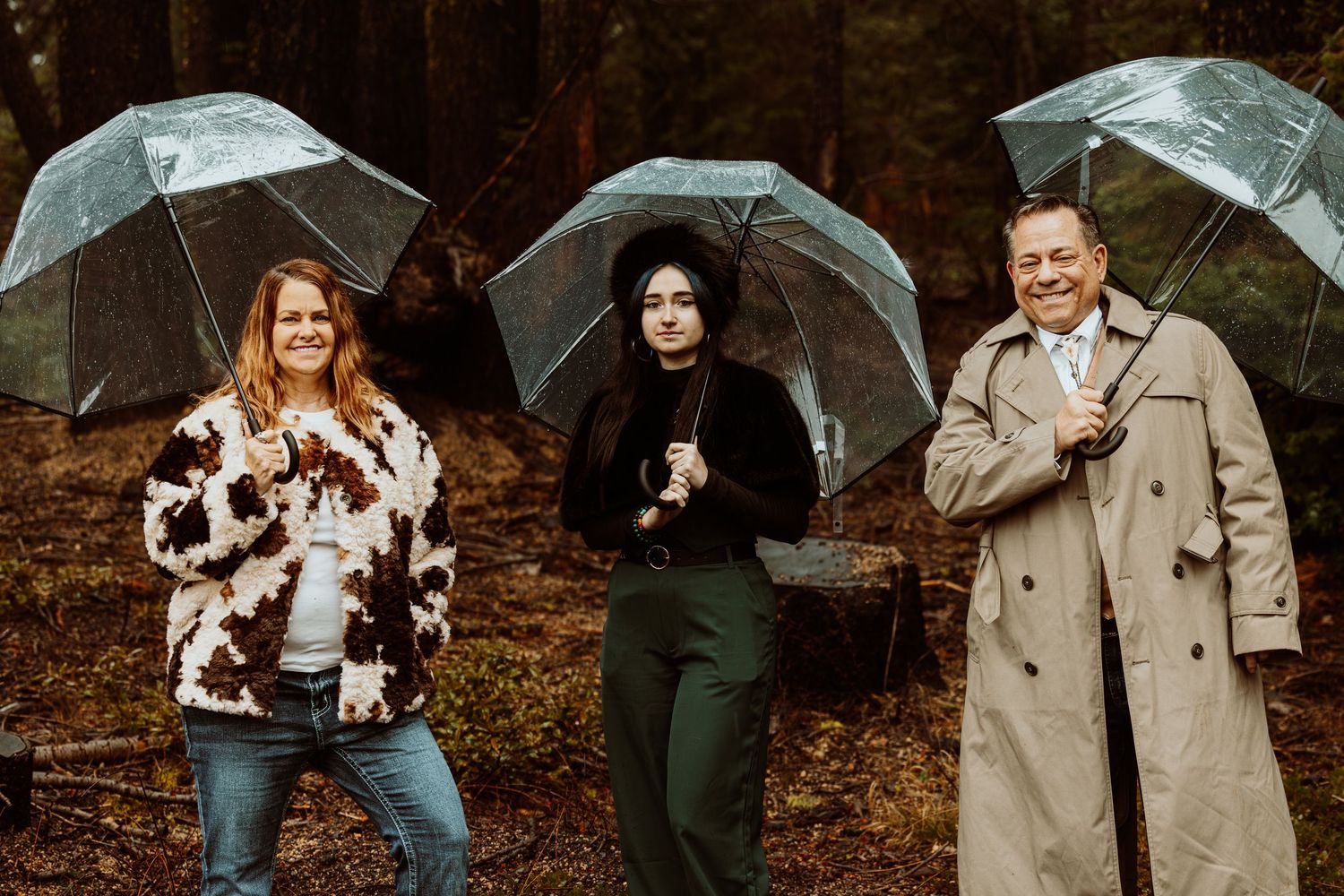 Three guests smile holding clear umbrellas in a rainy forest setting at an outdoor wedding event.