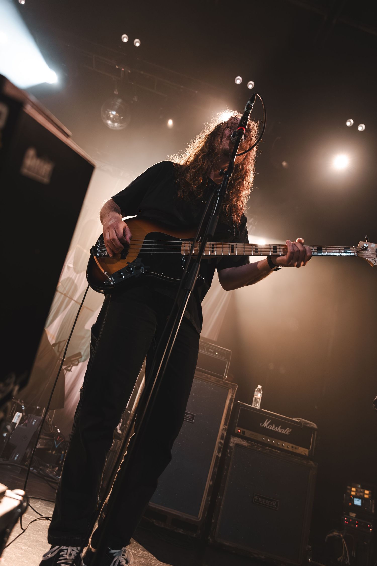 Long-haired bassist playing a sunburst bass guitar on stage surrounded by Marshall amplifiers.