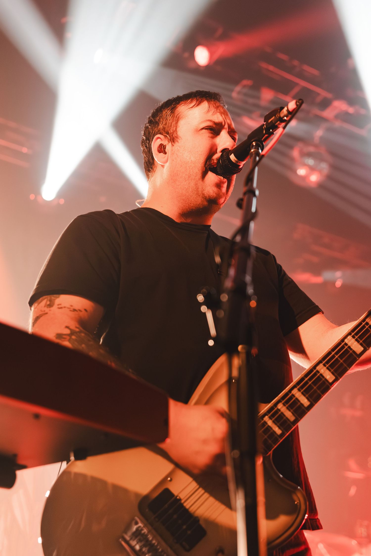 Tattooed rock musician sings and plays white guitar under dramatic red stage lighting at show.