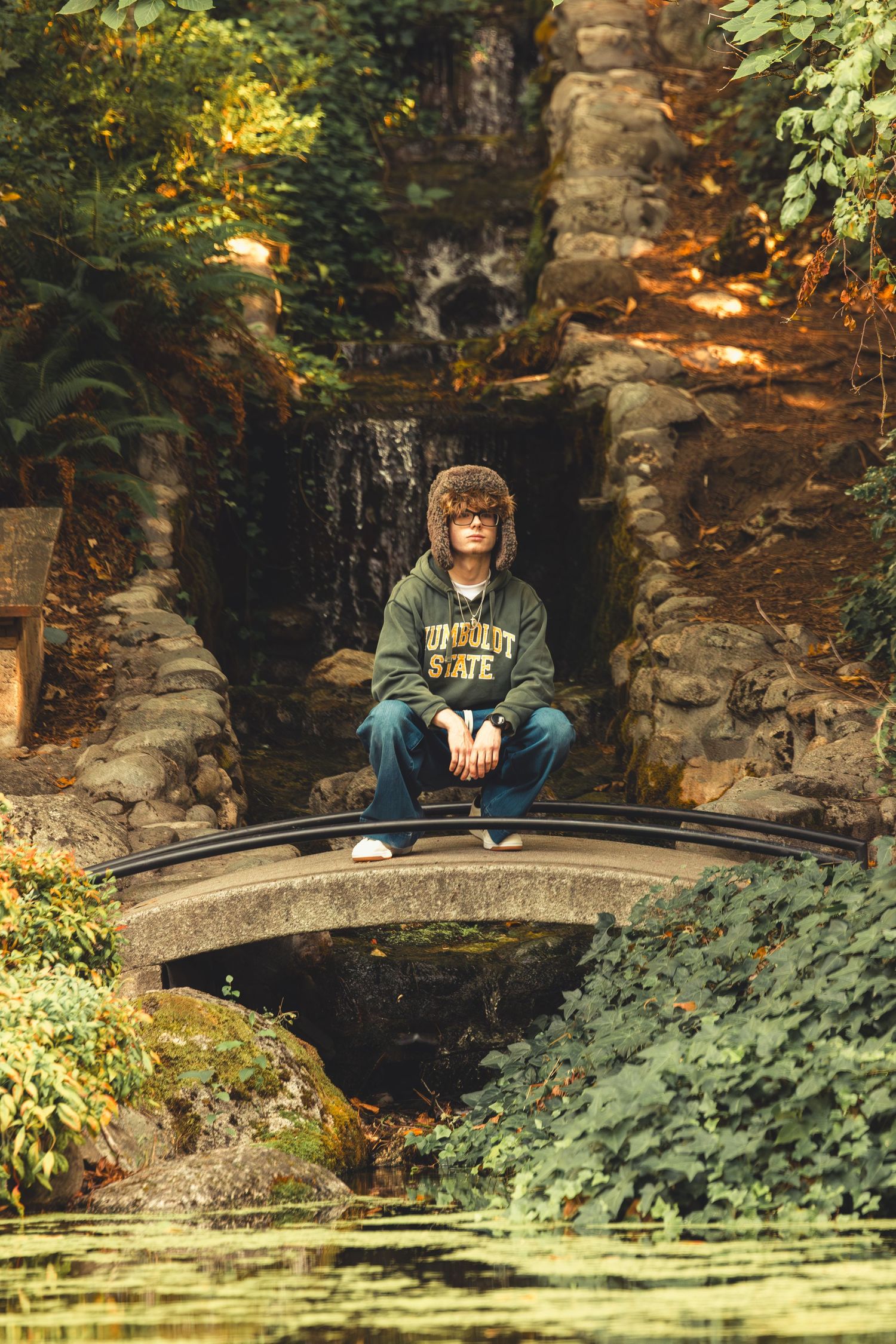 Teen in Humboldt State hoodie and fur hat sits cross-legged on stone bridge over pond in lush garden.