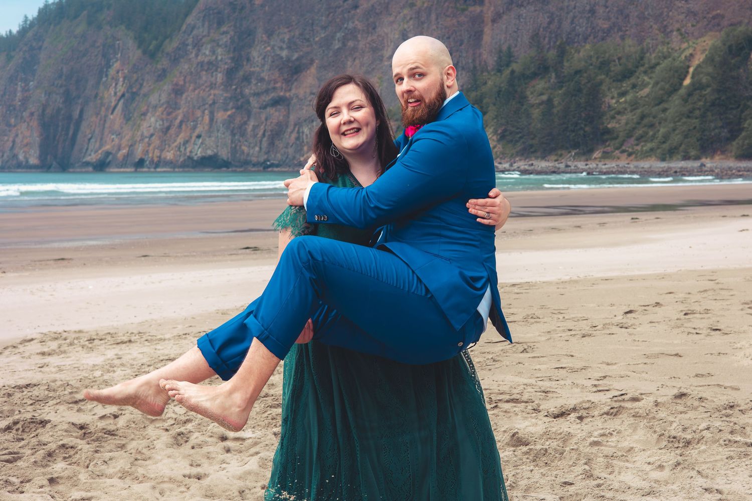 Groom in blue suit carries woman in green dress on a sandy beach with ocean backdrop.