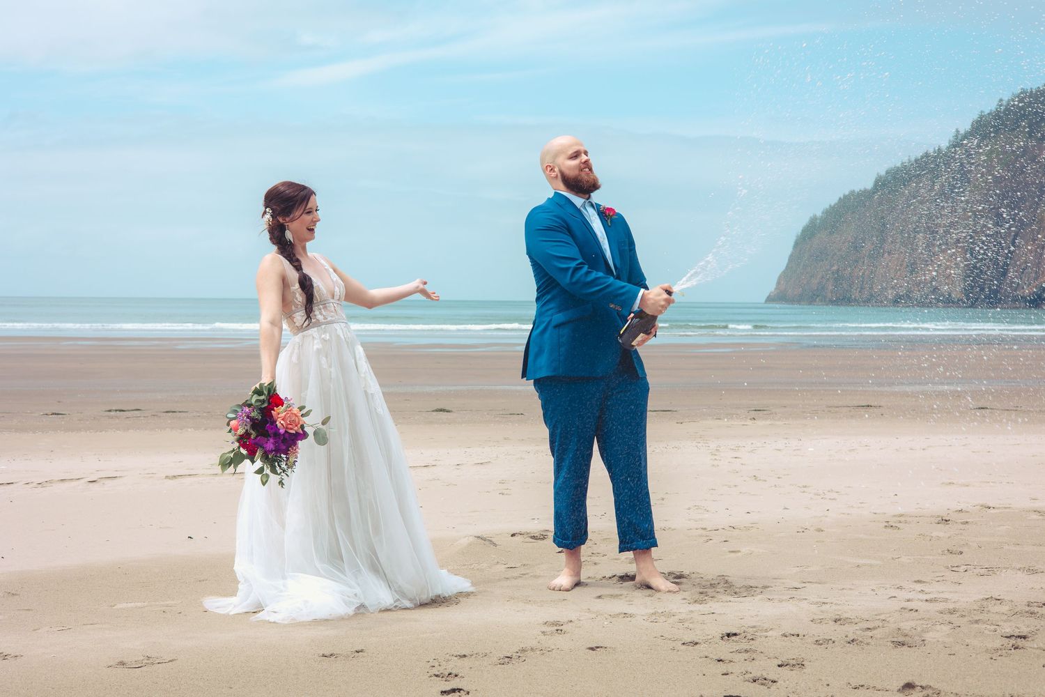 Bride and groom celebrating with champagne on a sunny beach with a rocky headland backdrop.
