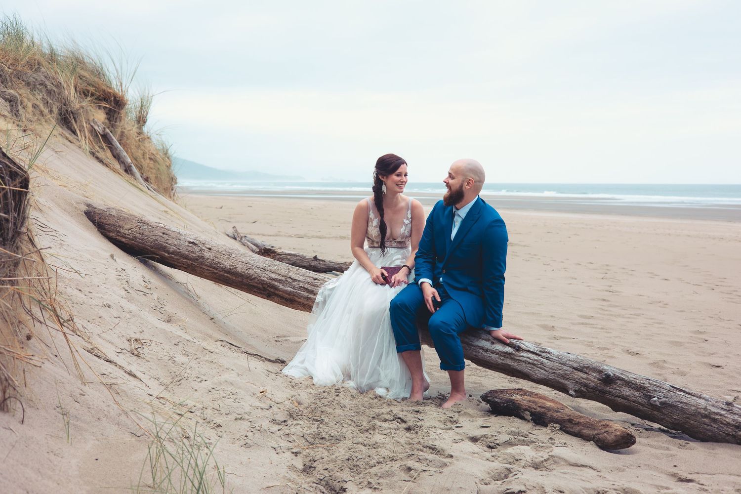 Bride and groom sit on driftwood logs smiling at each other on a sandy beach with dunes behind them.