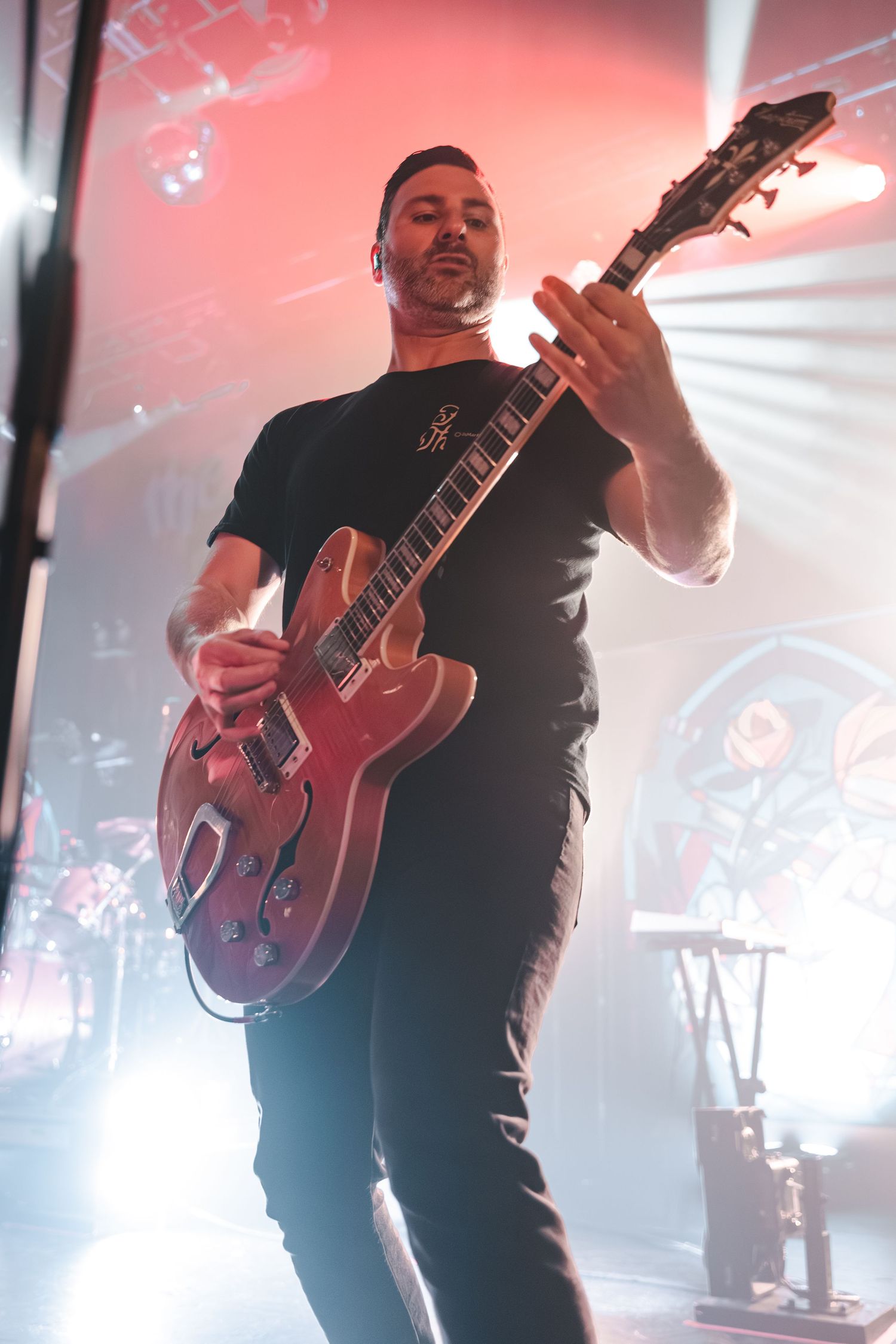 Guitarist playing red semi-hollow electric guitar on stage under dramatic lighting.