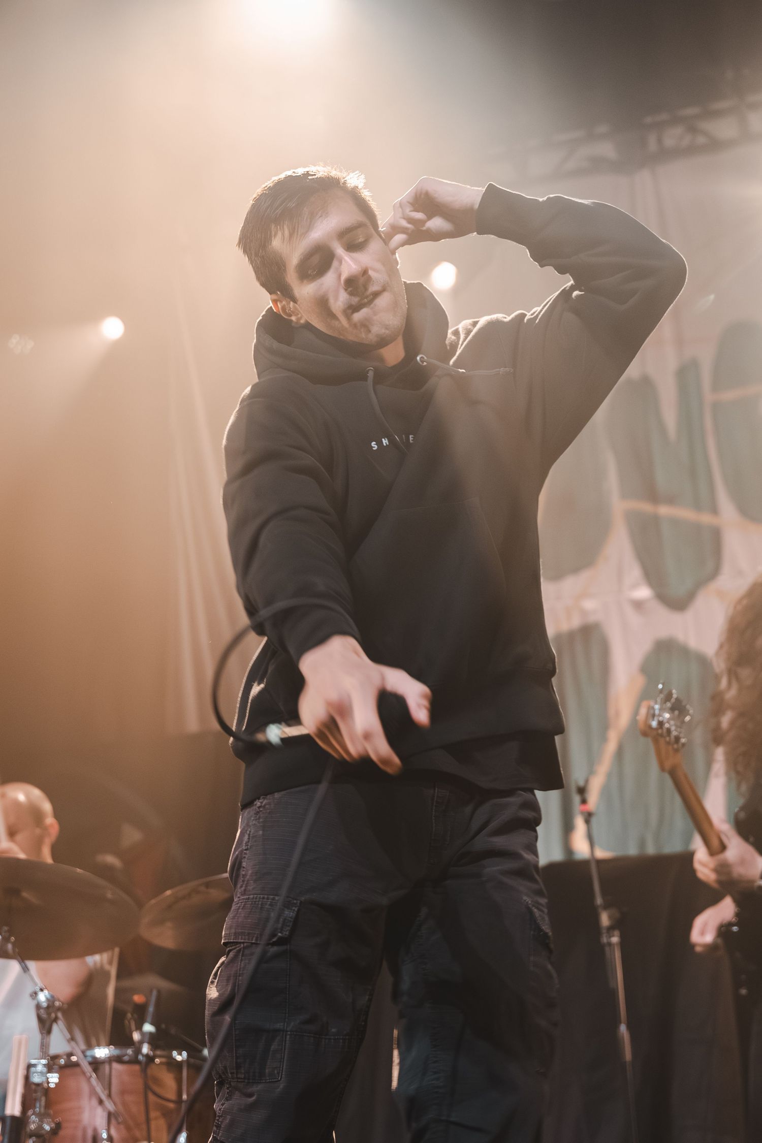 Male vocalist in black hoodie performing energetically on a lit concert stage holding a microphone.