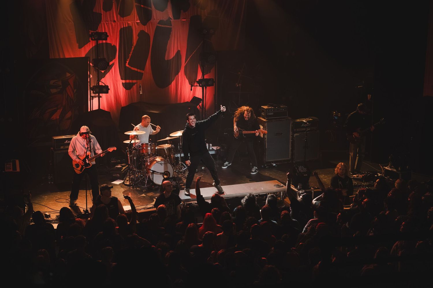Rock band performing on lit stage with abstract red backdrop art and audience silhouettes in foreground.