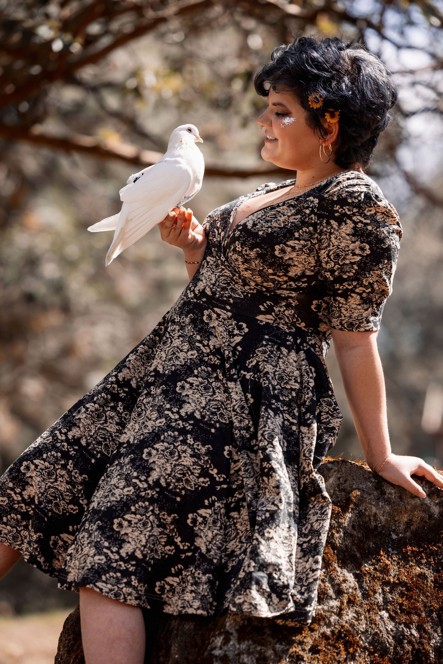 Woman in black floral dress gazes at a white dove perched on her hand while leaning against a sun-lit rock outdoors.