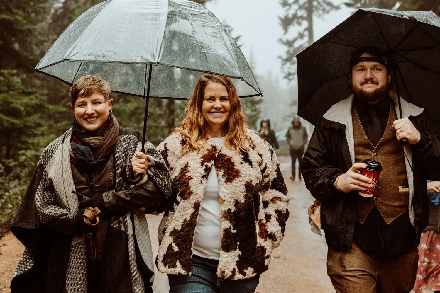 Three smiling friends walking on rainy forest path with umbrellas at outdoor event.