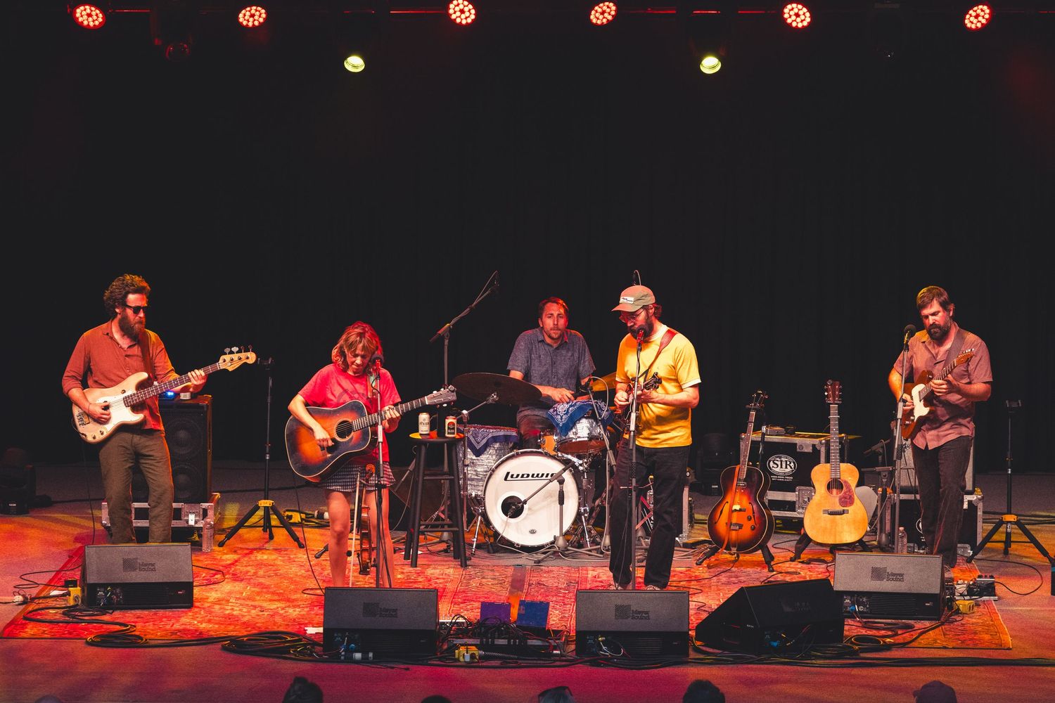 Five-piece band performing on a warmly lit indoor stage with guitars and drums before a live audience.