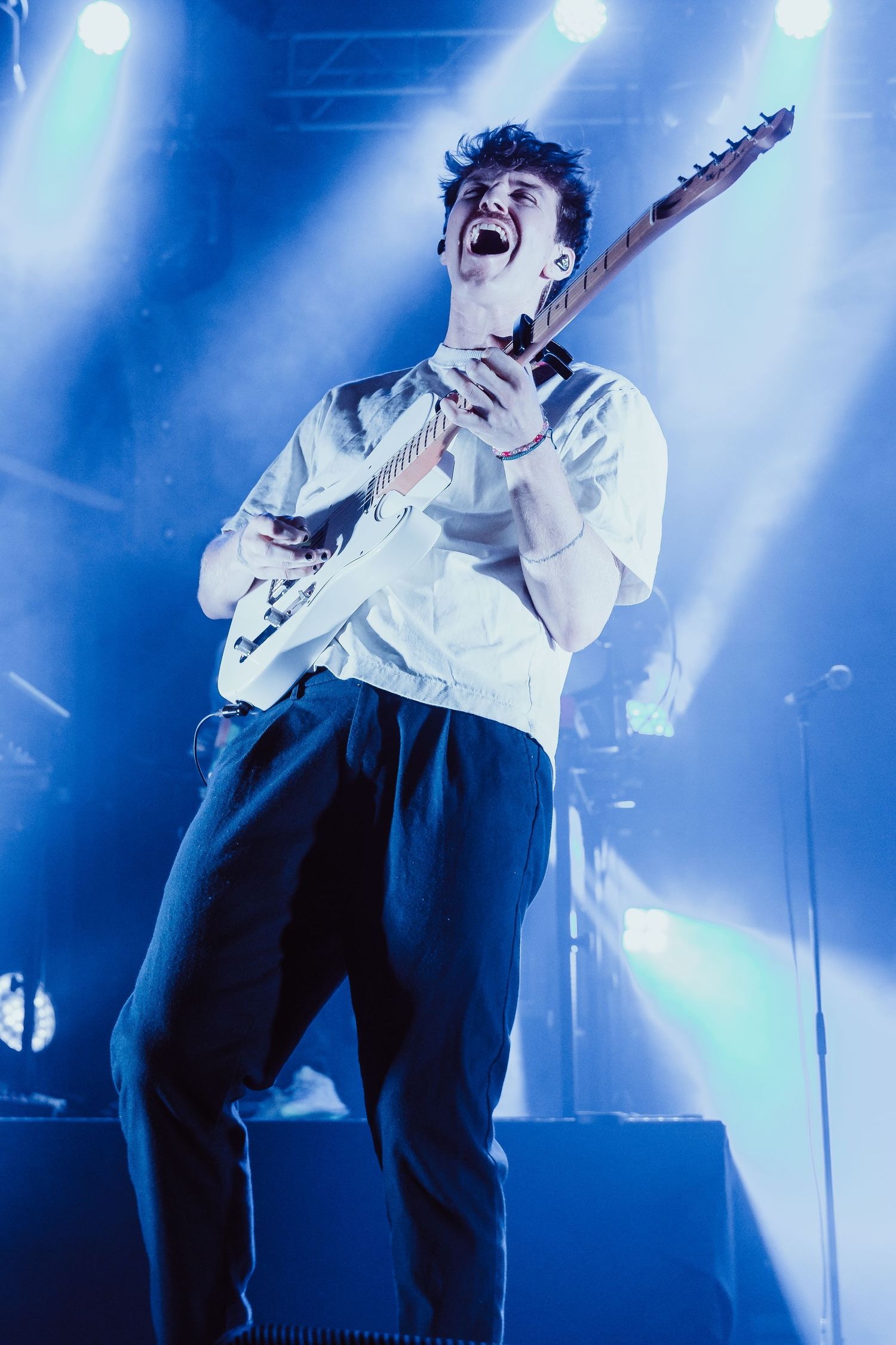 Young guitarist singing passionately while playing white electric guitar under blue stage lights.