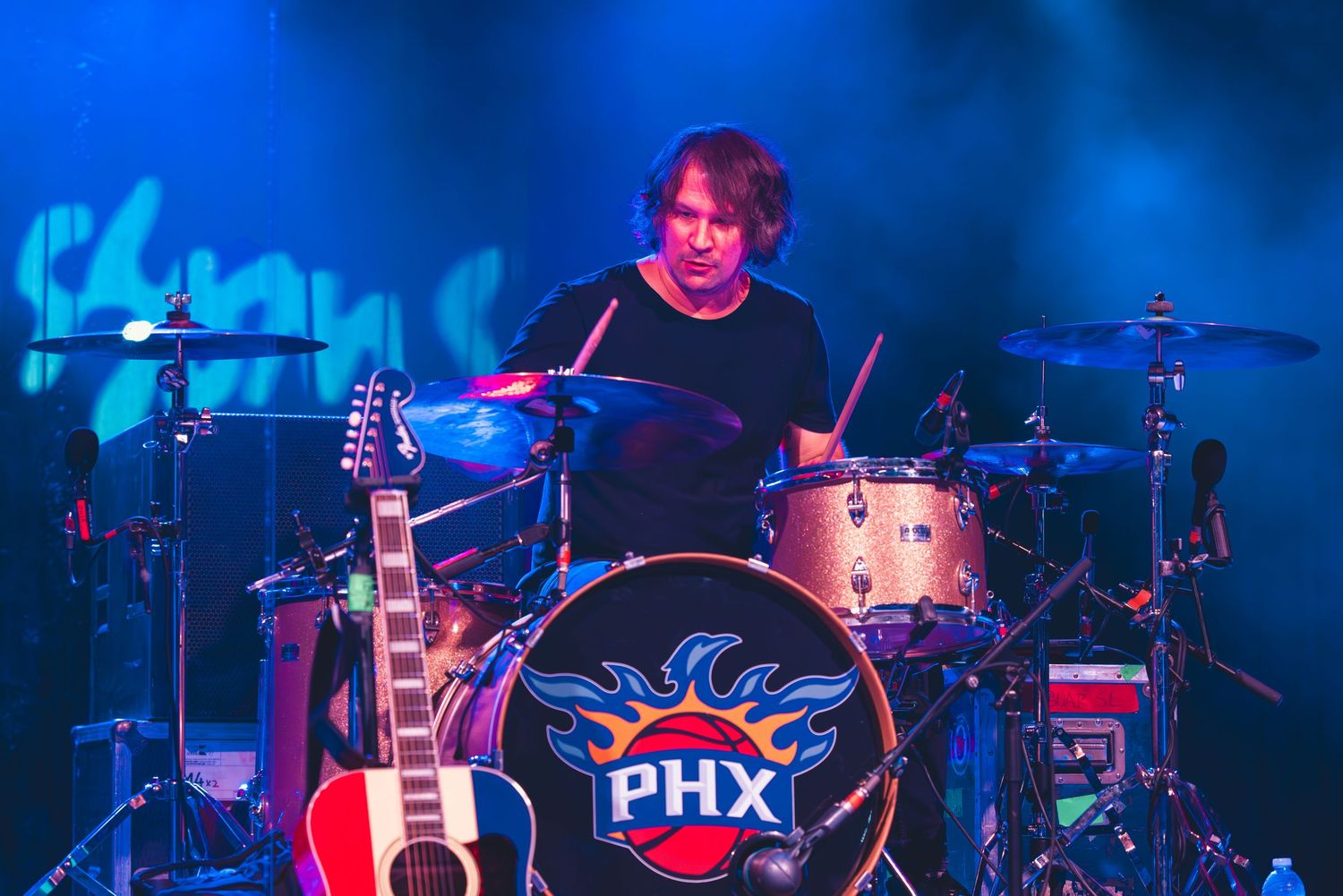 Drummer plays behind a kit featuring a Phoenix Suns PHX logo bass drum on a lit stage.