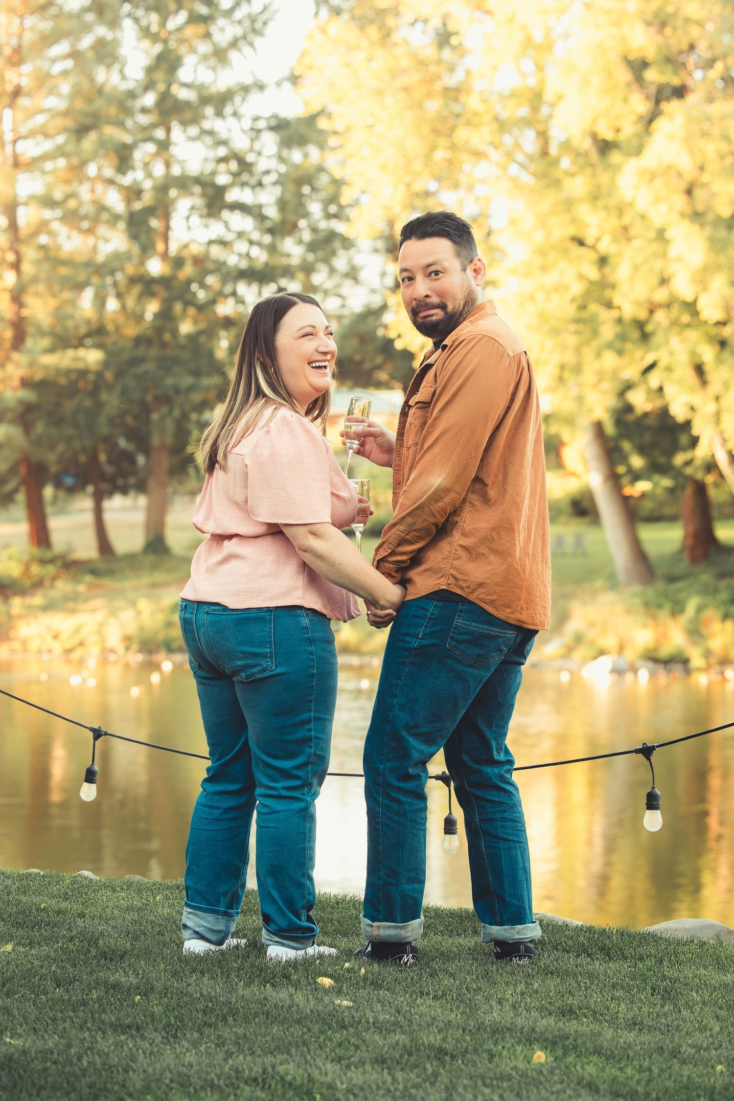 Couple toasting champagne glasses by a pond in an autumn park, smiling and holding hands.