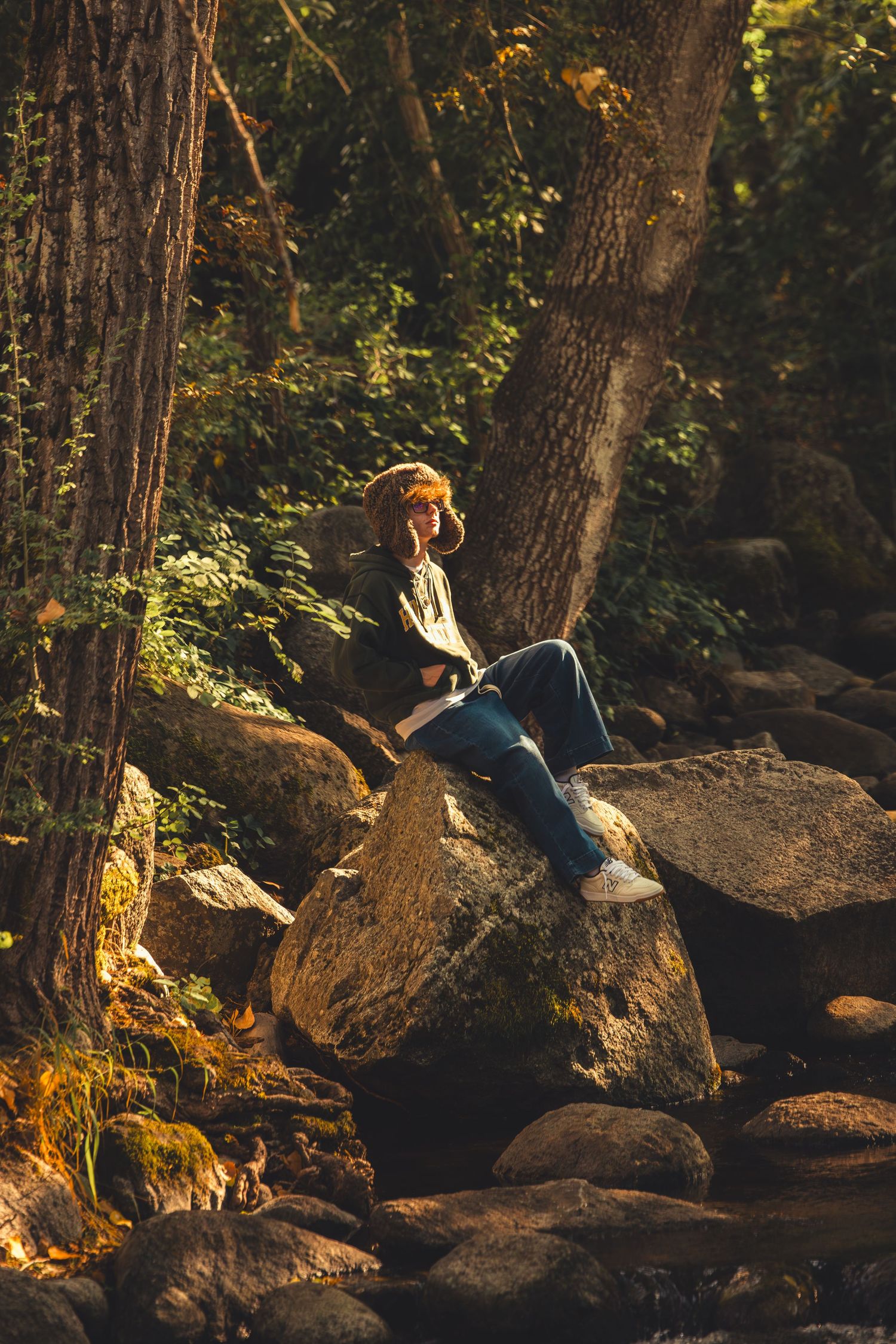 Redhead teen sits on mossy rocks by forest stream in golden autumn sunlight wearing jeans and hoodie.