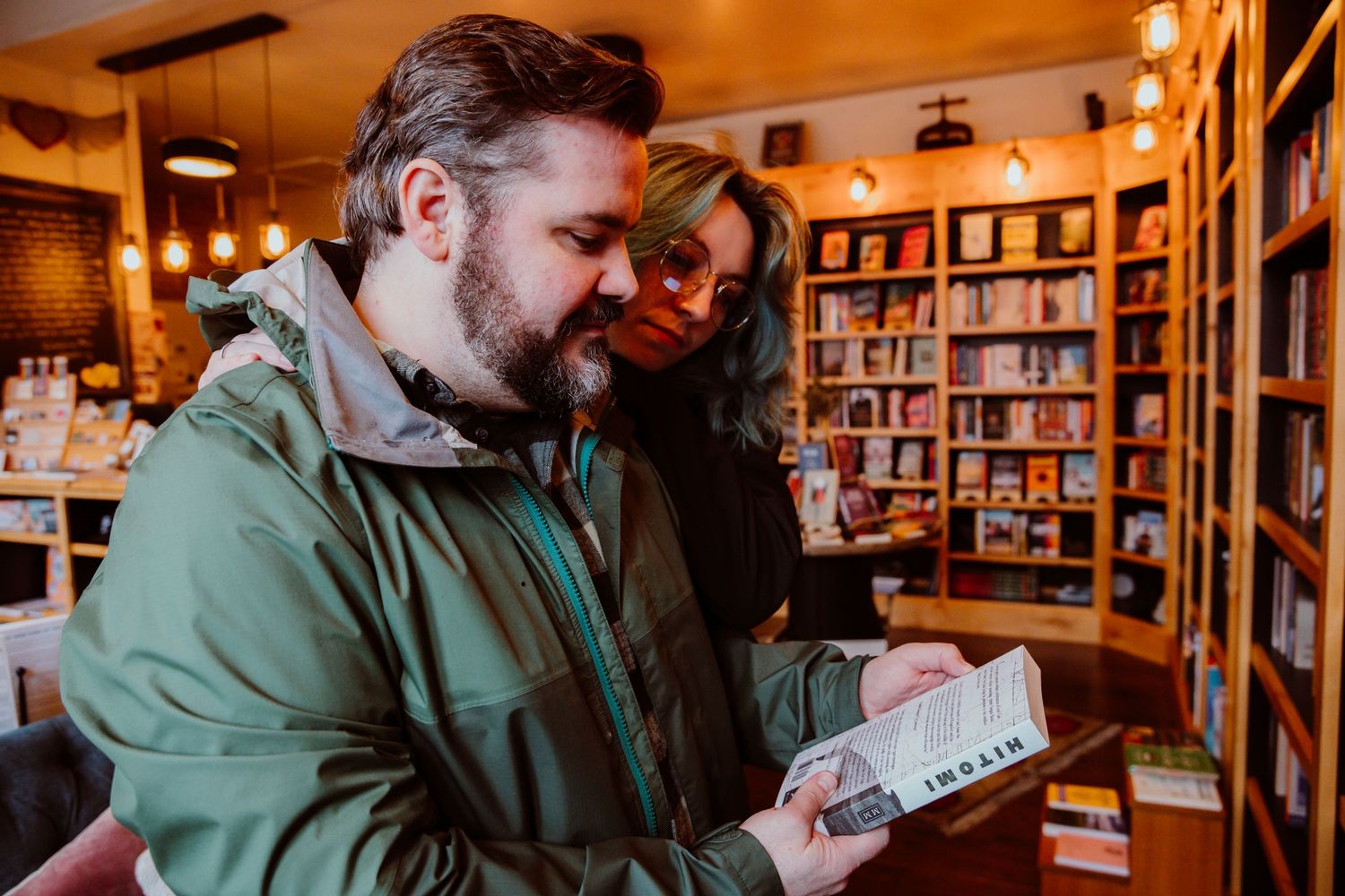 Bearded man in green jacket reading a book in a cozy bookstore with a woman behind him.