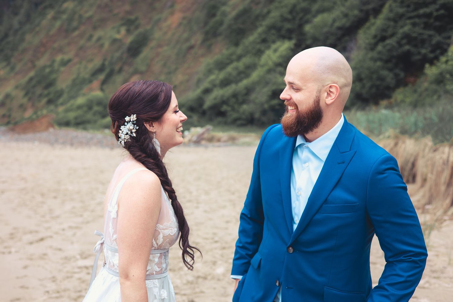 Bride and groom share a joyful moment on a sandy beach with green cliffs behind.