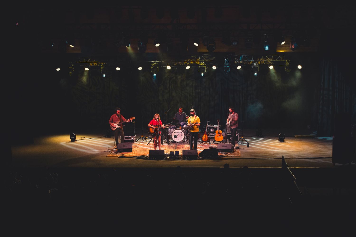 A five-piece band performs on a large concert stage under colorful stage lighting.