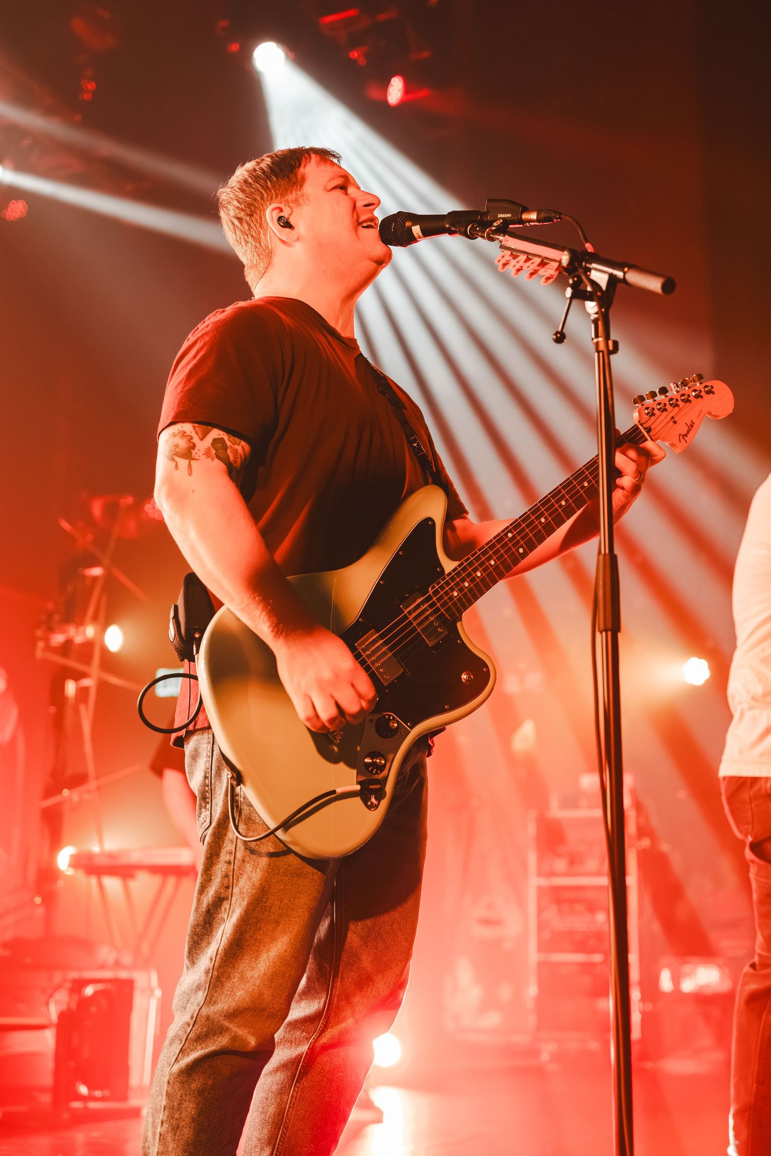 Rock guitarist sings passionately into microphone while playing white guitar under red stage lights.