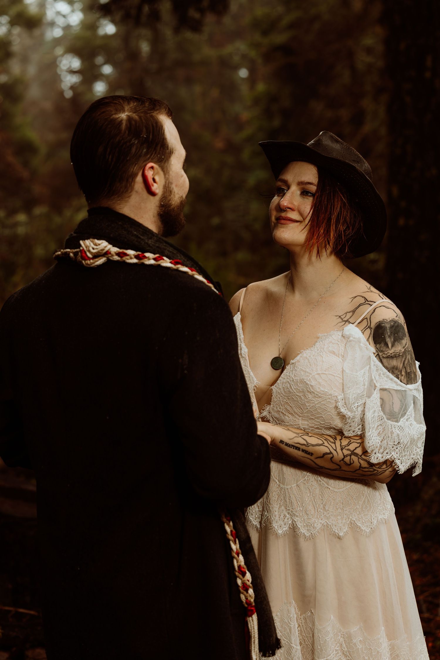 Tattooed bride in white lace dress and black hat holding hands with groom in dark forest.