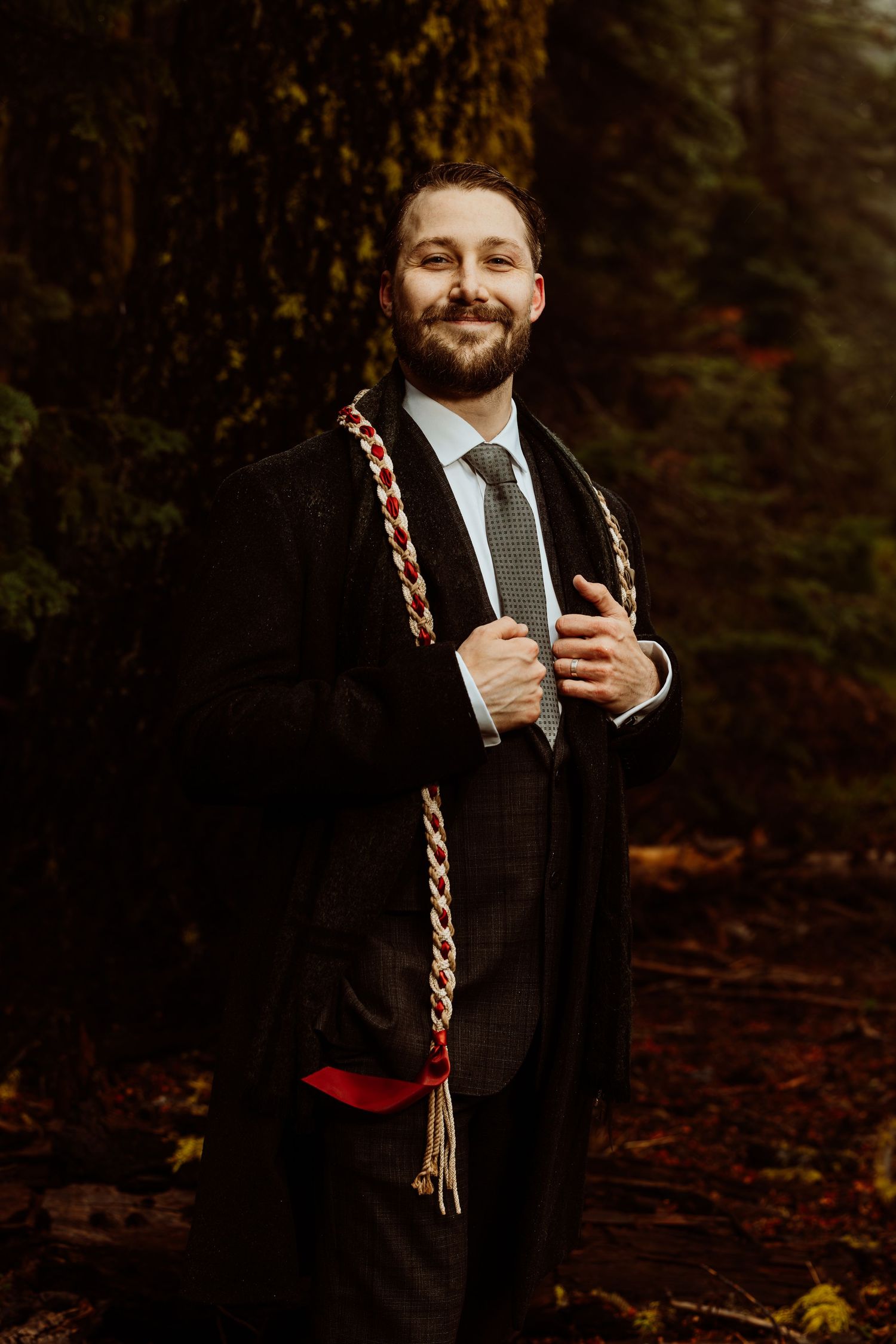 Smiling bearded groom in dark suit with colorful braided ceremonial sash, standing in forest.