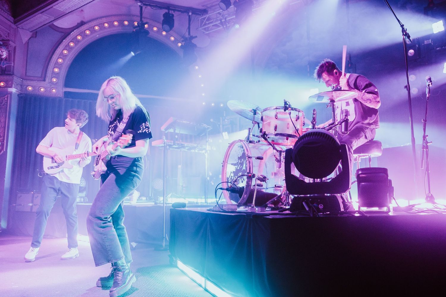 Rock band performing on a purple-lit stage in an ornate venue with three musicians.