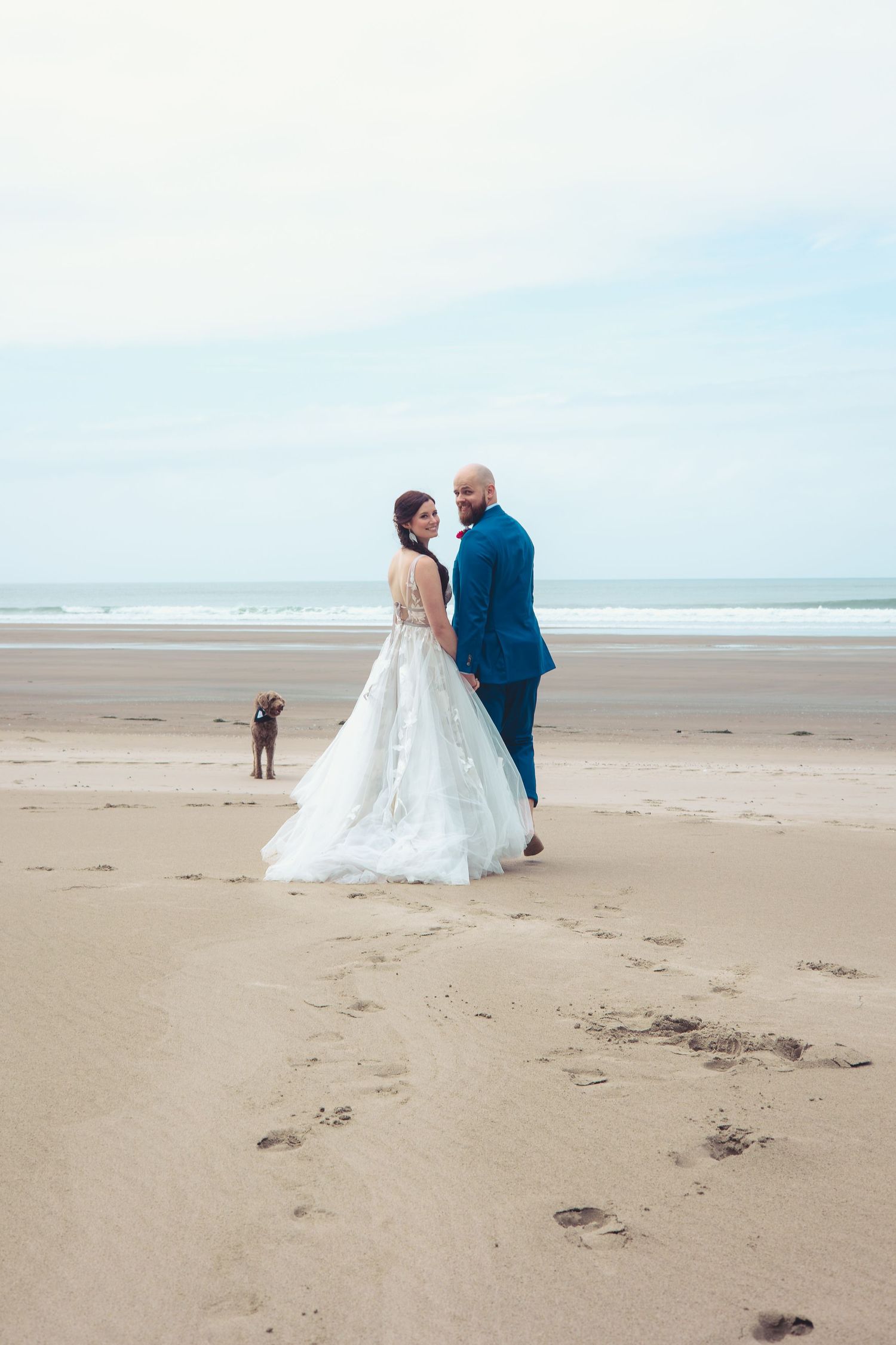Bride in white gown and bald groom in blue suit pose on sandy beach with dog.