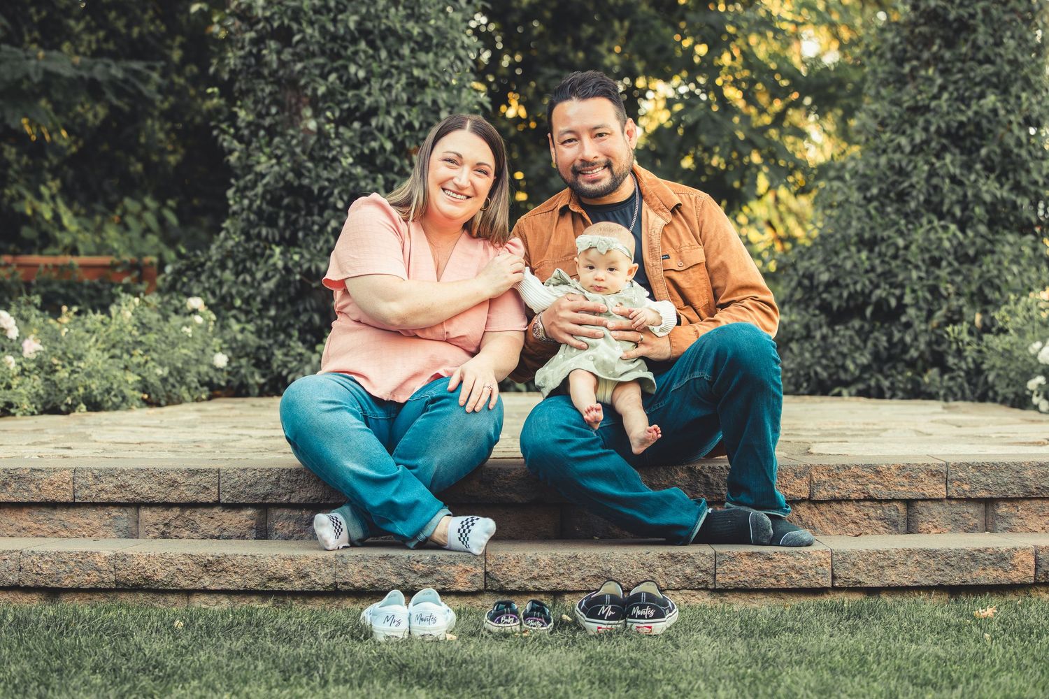 Happy family of three sitting outdoors on stone steps with baby shoes displayed in front, surrounded by lush greenery.