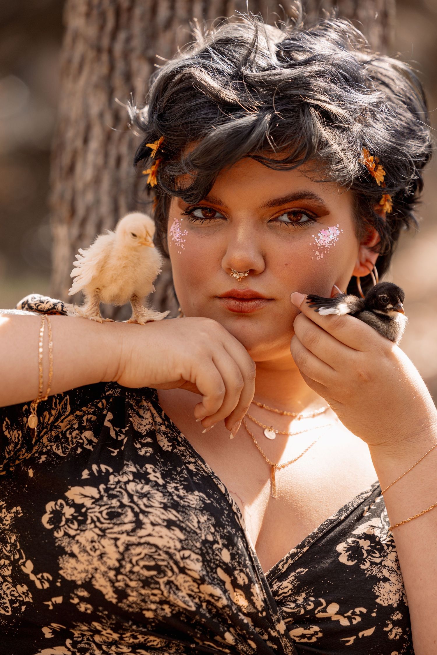 Woman with glitter makeup and nose ring poses with two baby chicks perched on her shoulder and hand.