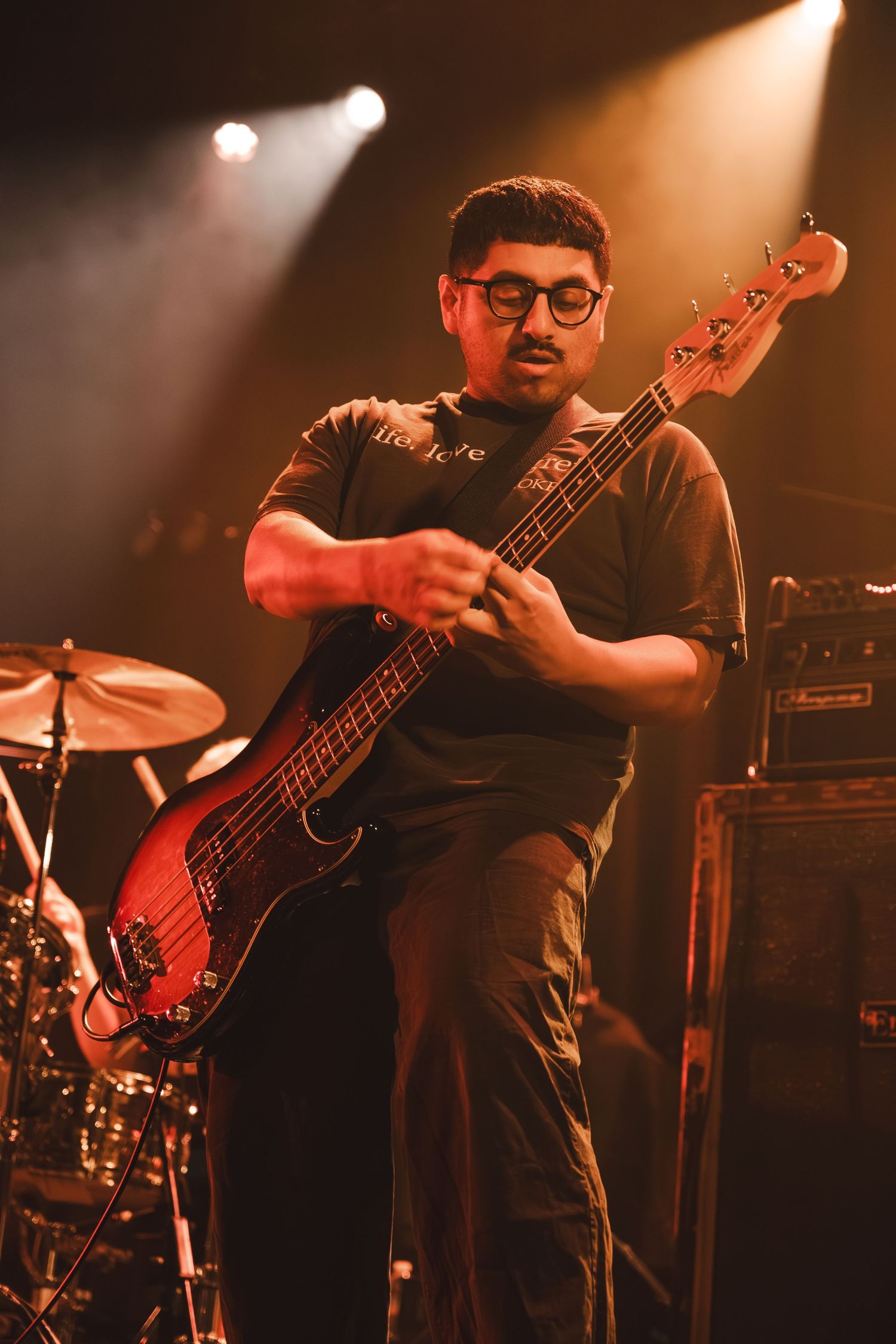 Close-up of male bassist with glasses intensely playing red bass guitar under warm stage lights.