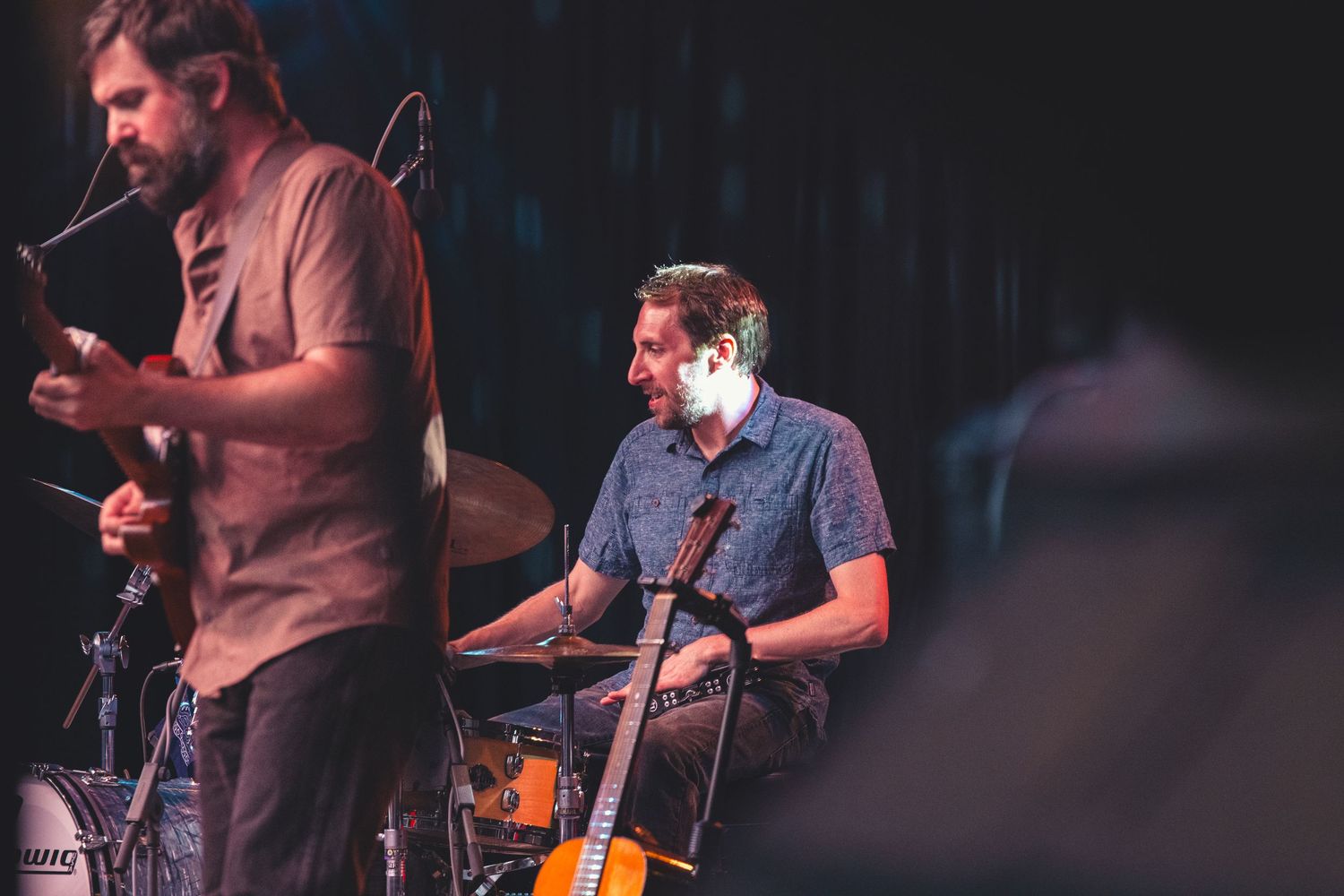 Guitarist and drummer performing together on a moody, dimly lit concert stage.
