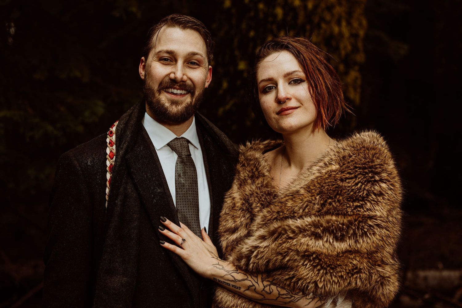 Bride in fur stole and groom in dark suit smiling together in an autumn forest wedding photo.